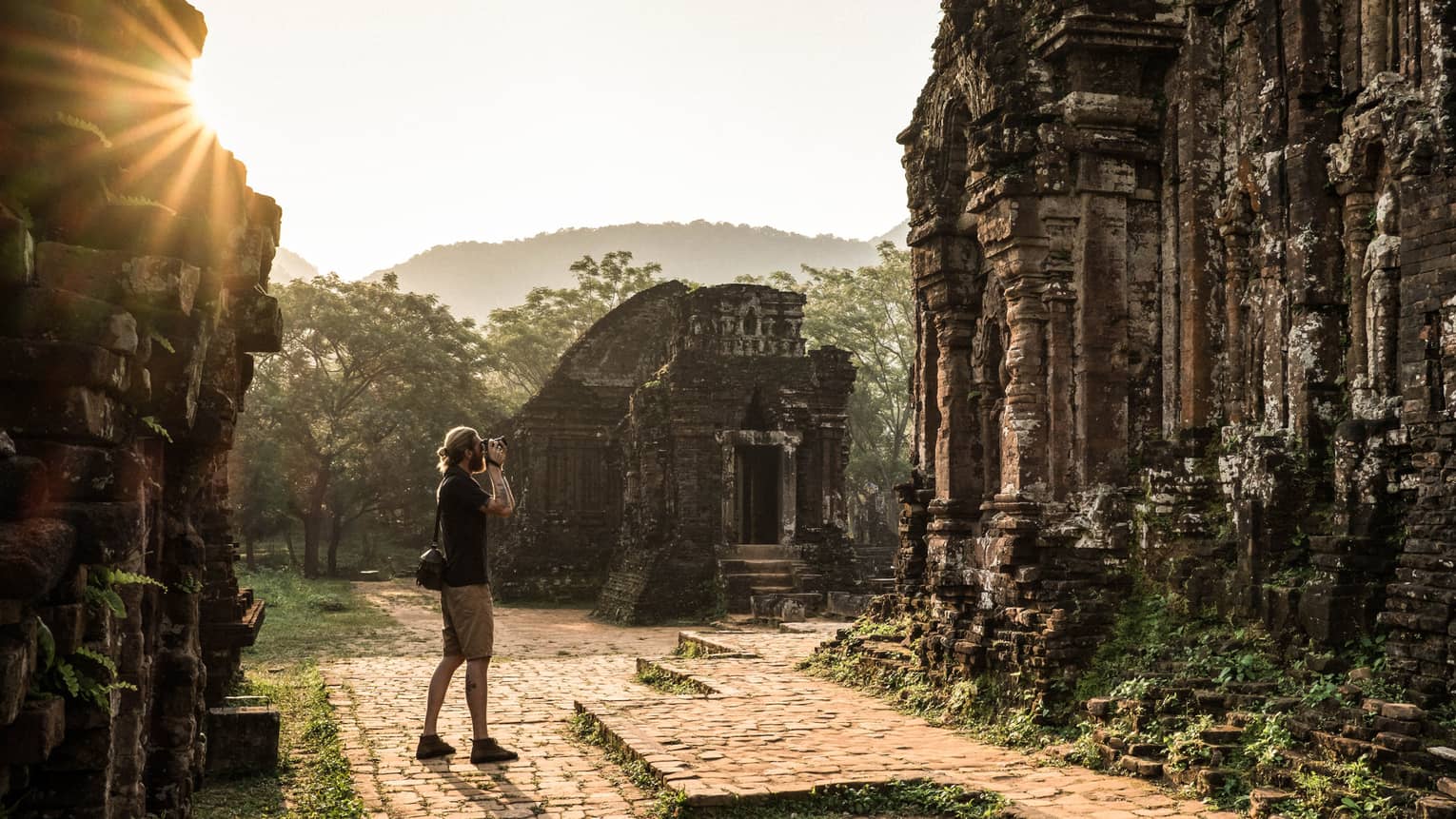 A tourist photographing ancient stone ruins, surrounded by lush greenery and misty hills in the background.