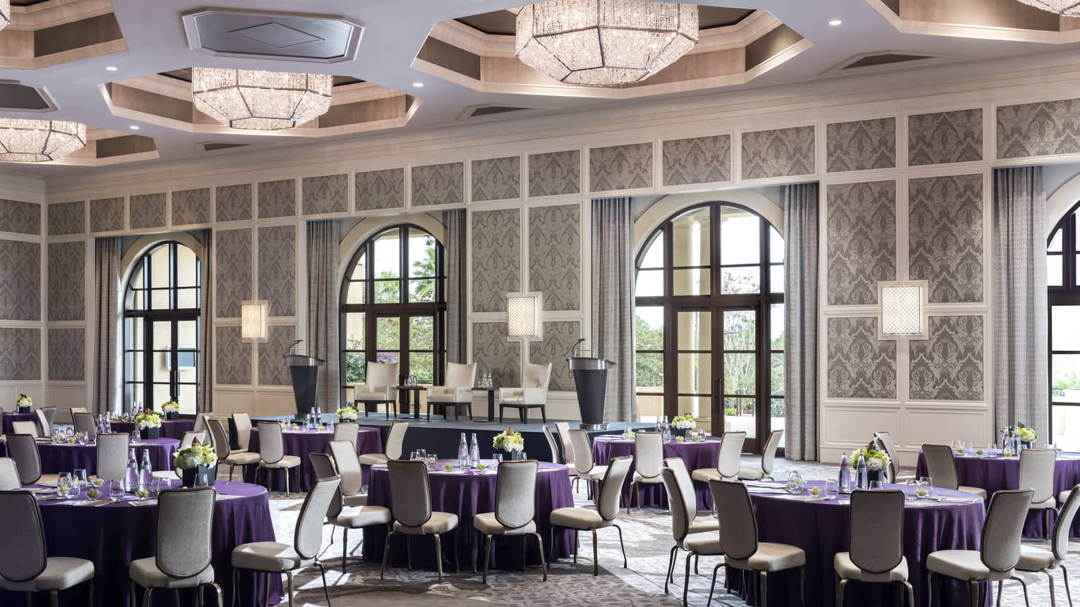 Ballroom with round banquet tables under geometric chandeliers, sunny arched windows 