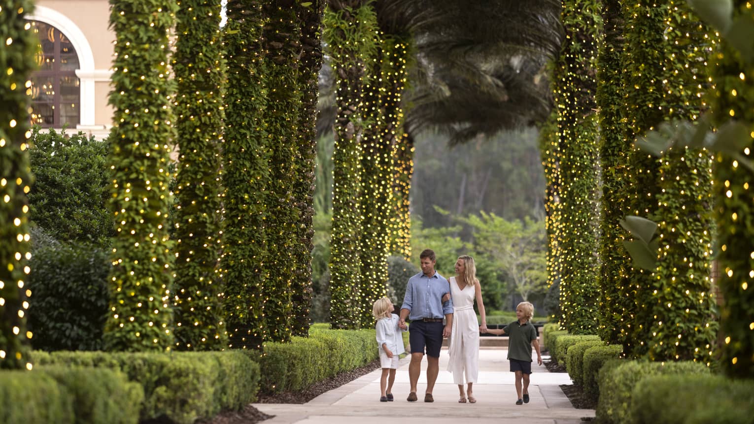A mother, father and two sons walking along a path of palm trees wrapped in Christmas lights.