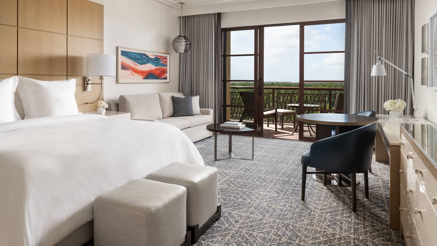 Hotel room with white bed, beige sofa, small table and two black chairs, walk-out balcony