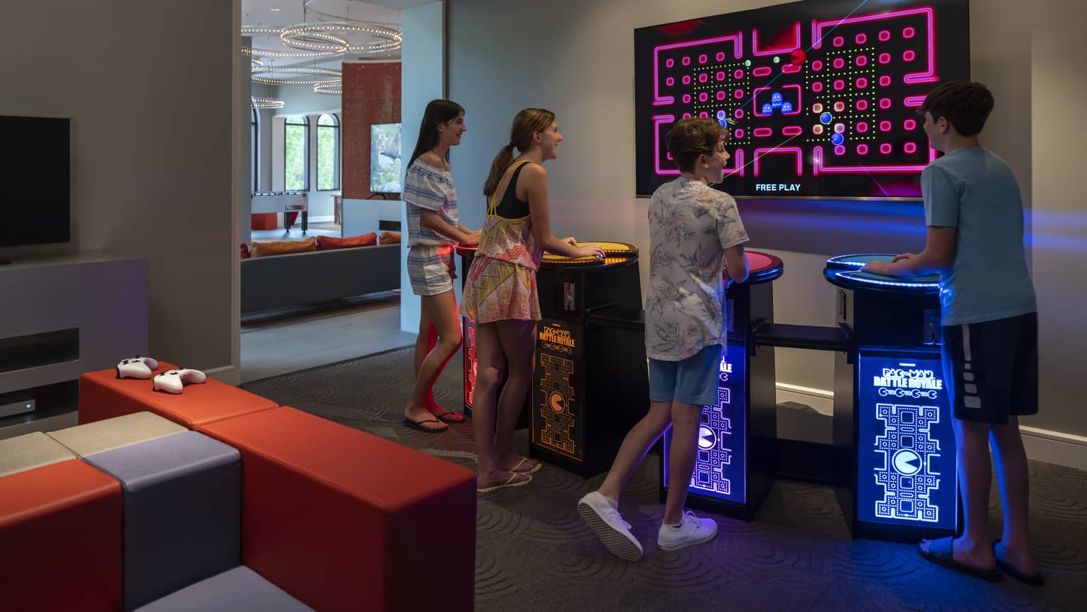 Four smiling teenagers stand at multi-coloured video-game pedestals facing a large TV screen in a dimly-lit games room.
