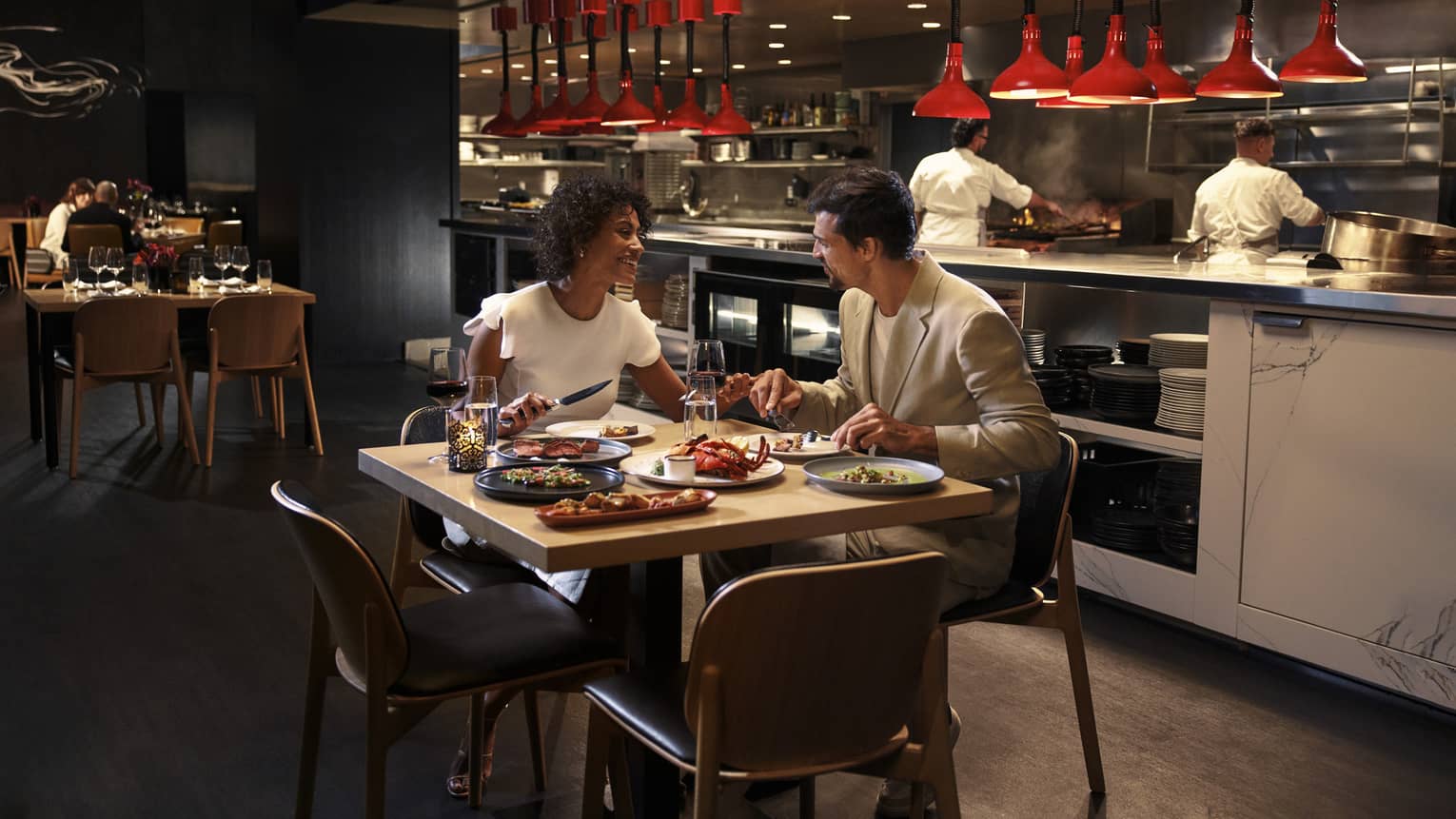 Two people sit at a square dining table in front of an open kitchen with two chefs working behind them