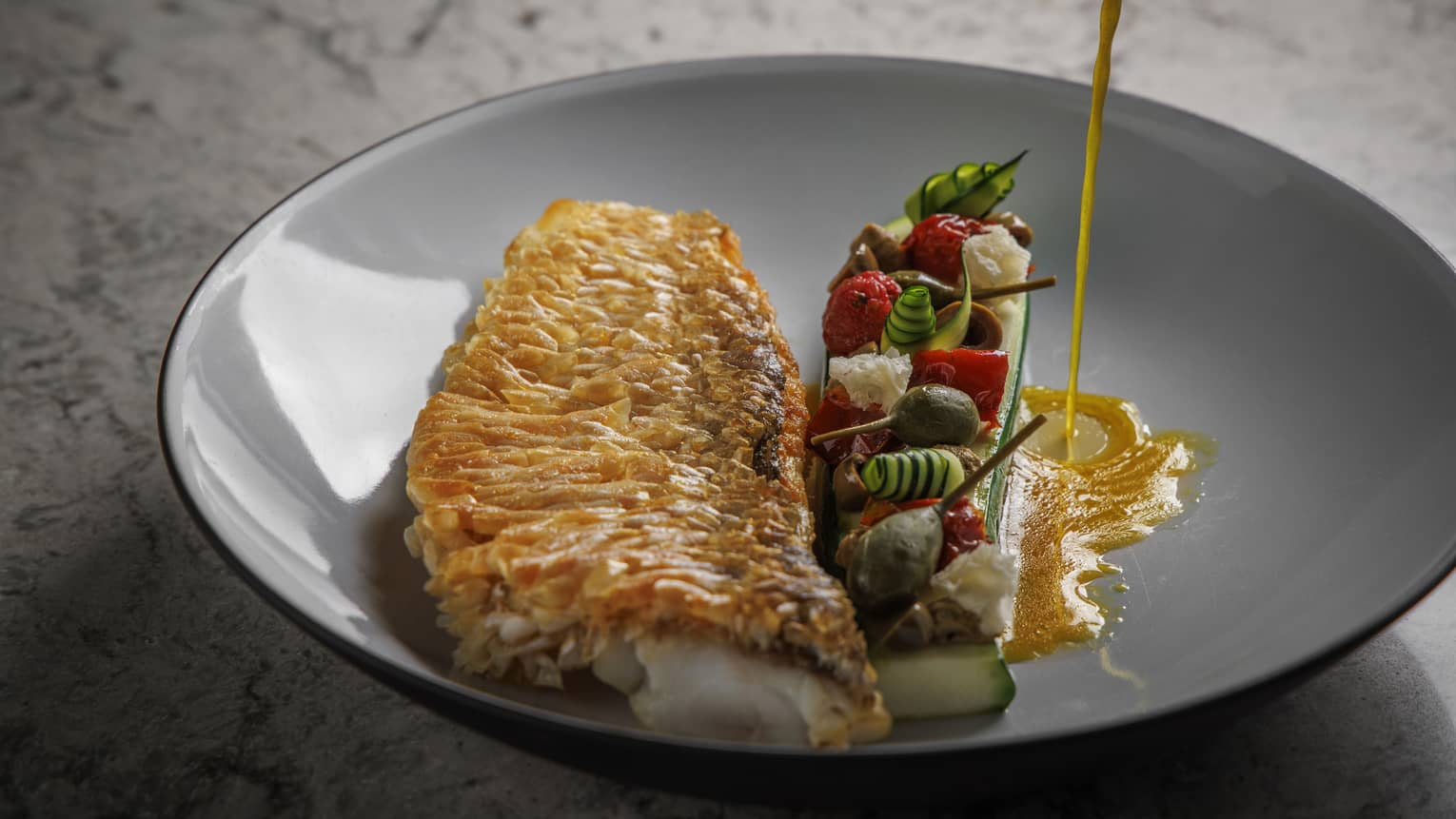 Fish filet with assorted vegetables in shallow grey bowl, sauce being poured into it