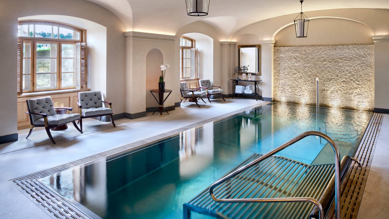 Blue indoor swimming pool, sunny windows, lanterns hanging from ceiling, deck with blue cushioned pool chairs