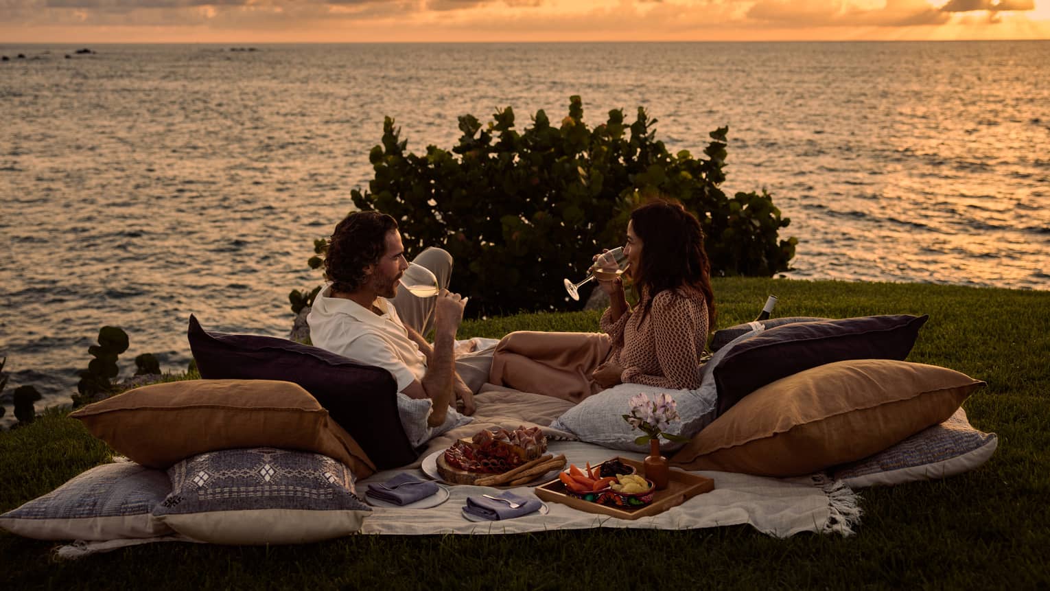 A man and woman lay on blanket and pillows during luxury picnic at sunset, next to the ocean