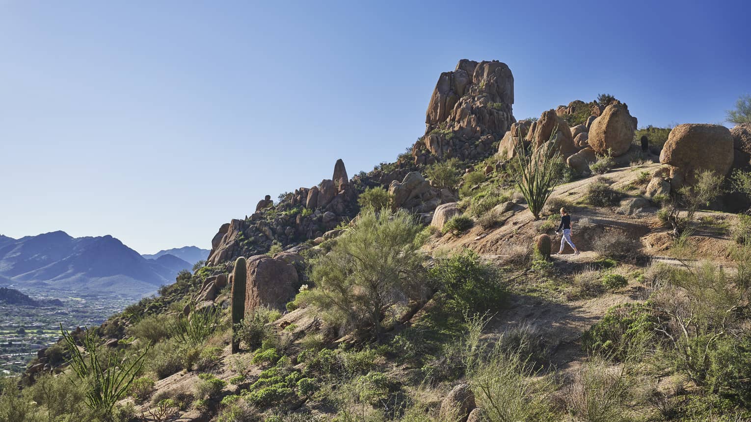 Woman walks along trail on rocky desert mountain under blue sky