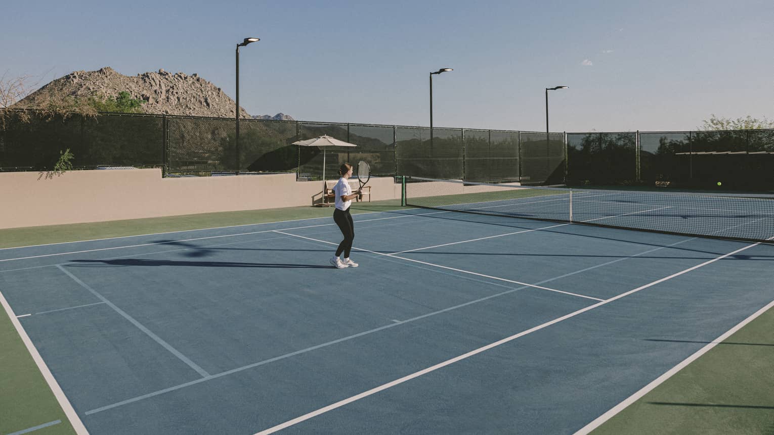 Person standing on a tennis court holding a tennis racquet with mountains in the background.