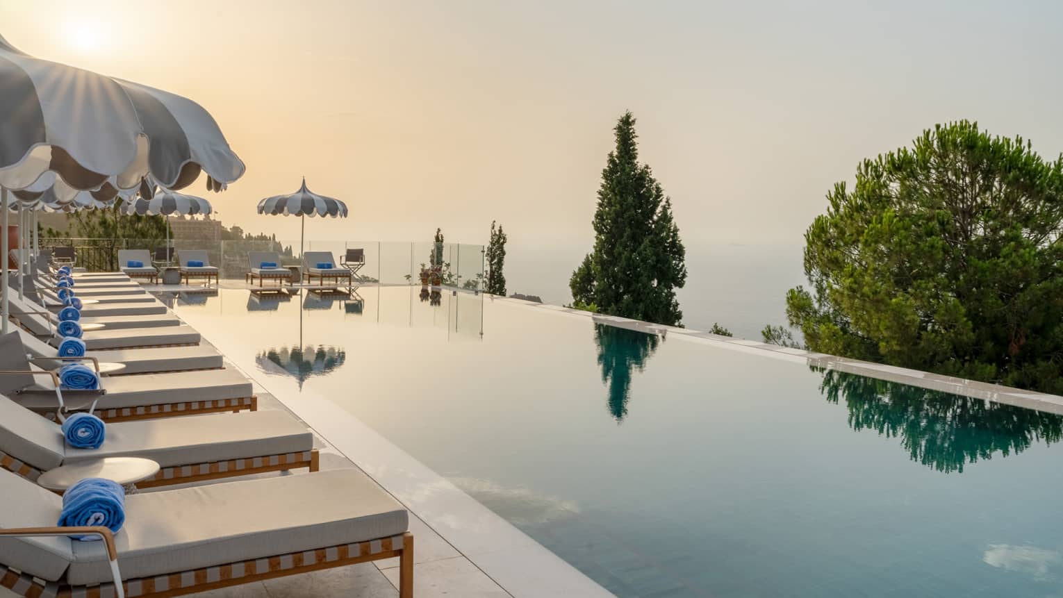 A long, smooth infinity-edge pool reflecting low sun on a partially cloudy day. Lounge chairs and umbrellas line one side and end of the pool, and tree tops and ocean views are in the background.