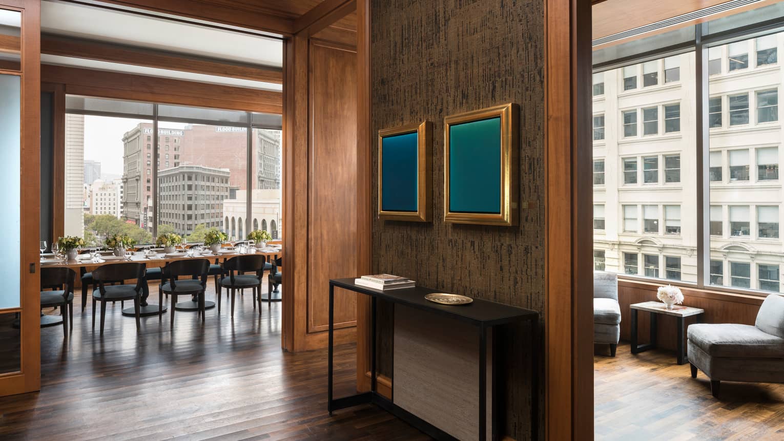 A waiting area in a restaurant with grey chairs and a small table, a private dining area with large windows can be seen through a hall.