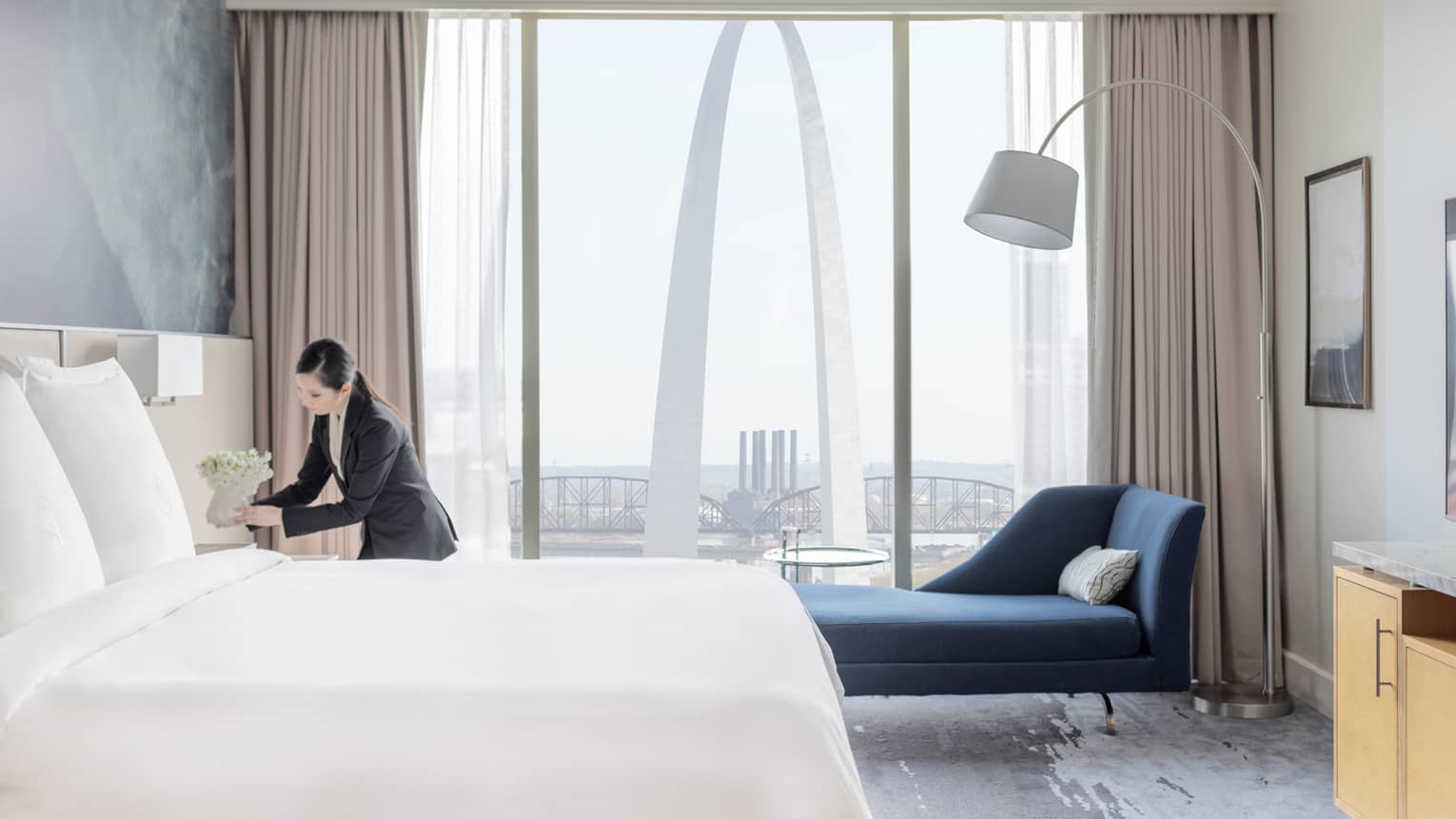 Hotel room with a king bed and blue chaise lounge by large windows with a view of the Gateway Arch, and a staff member arranging flowers on the nightstand