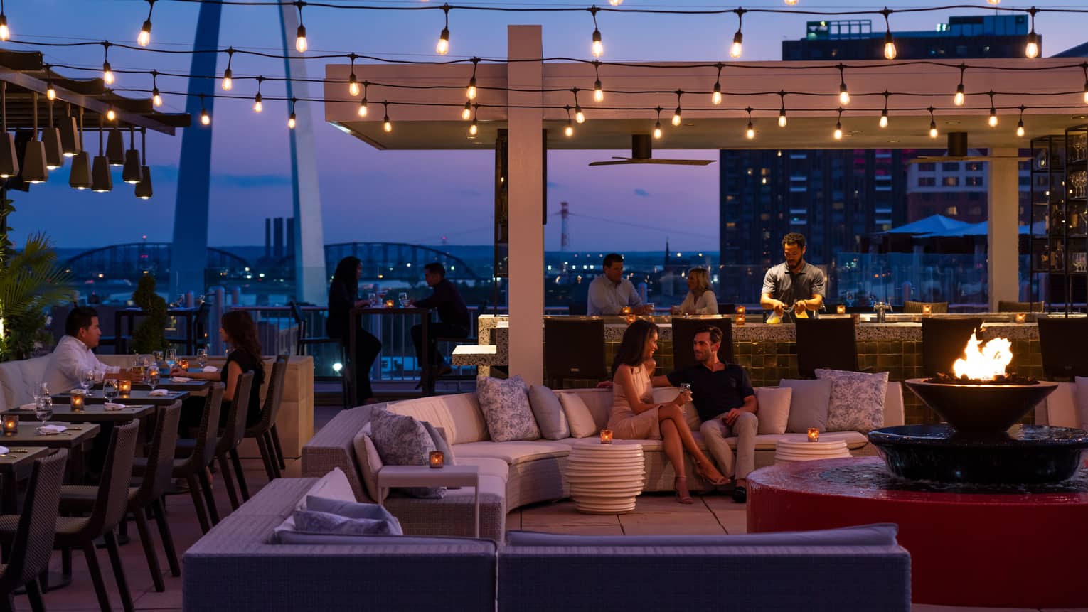 Rooftop bar and lounge with guests enjoying drinks and dining, featuring string lights, fire pit seating and a view of the Gateway Arch against the evening sky