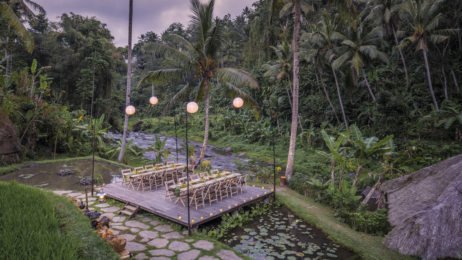 Two long tables laid out with food sit on a lantern-lit platform above a lotus pond and beside a river and dense forest.