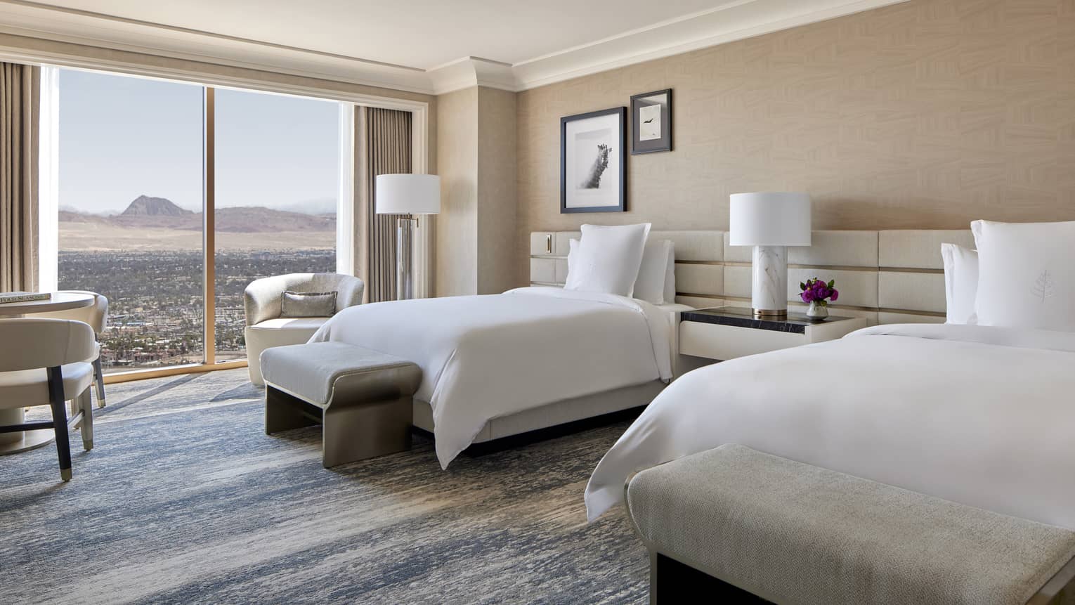 A room with two beds, large window, side tables, and chairs.