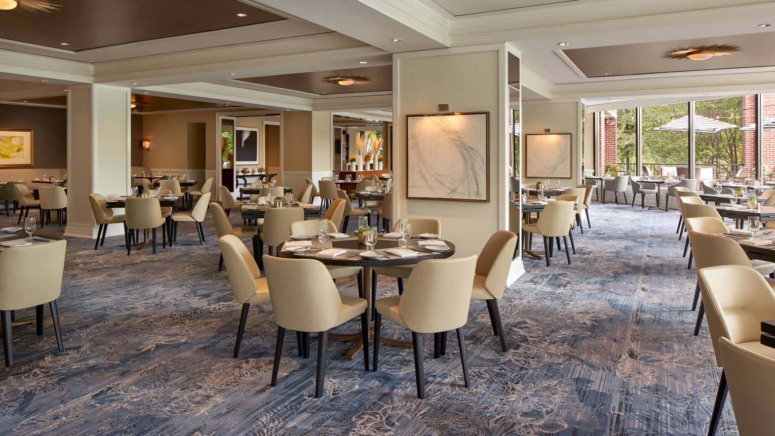 A well lit restaurant with beige chairs and round tables and large open windows.