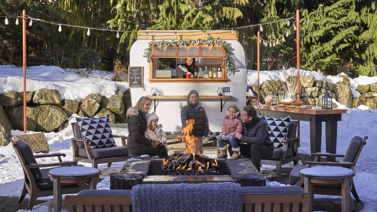 An outdoor camper sits behind a young family as they talk around a fire.