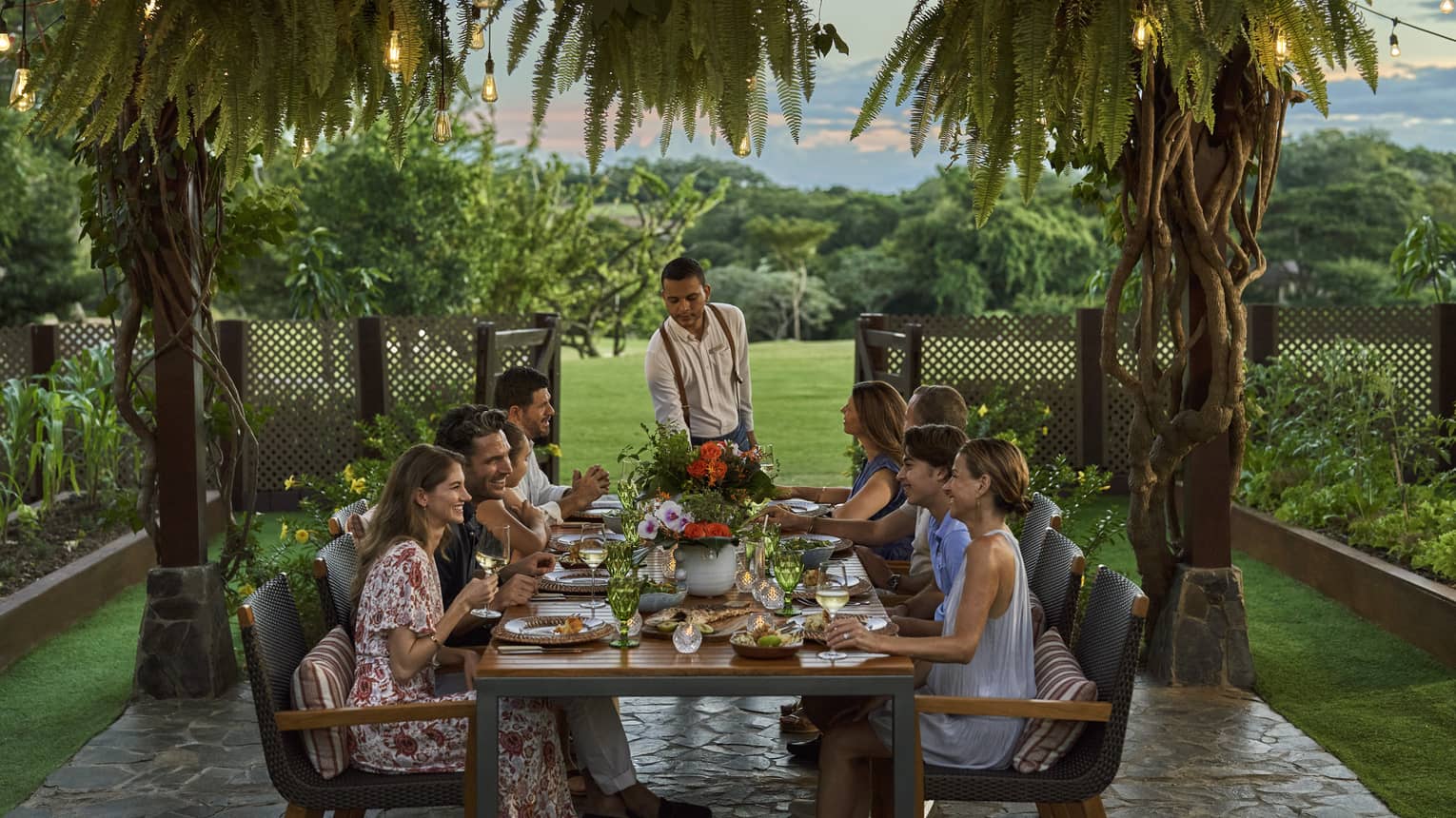 A group of people settled around a table, underneath a series of string lights and palm trees.