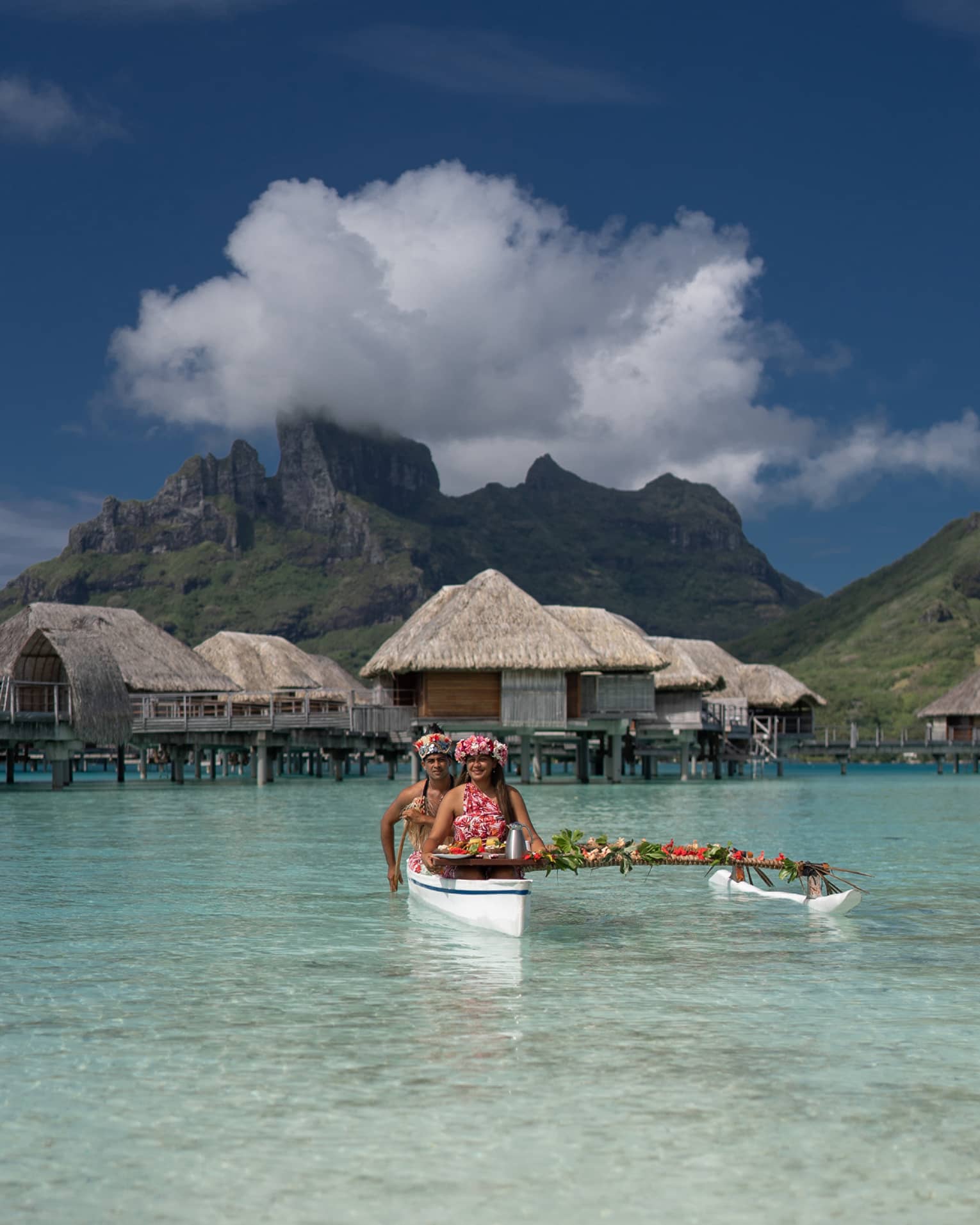 Two vibrantly dressed people in a canoe proffer a tray with colourful food attached to a pole draped with leaves and flowers.