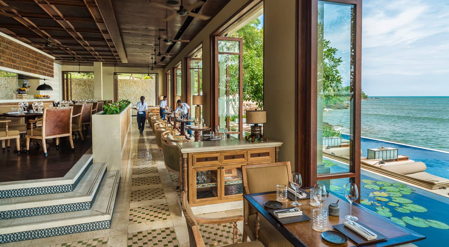 Airy dining room with tables, stairs down to cocktail tables by open floor-to-ceiling windows looking at lotus pond, pool, ocean