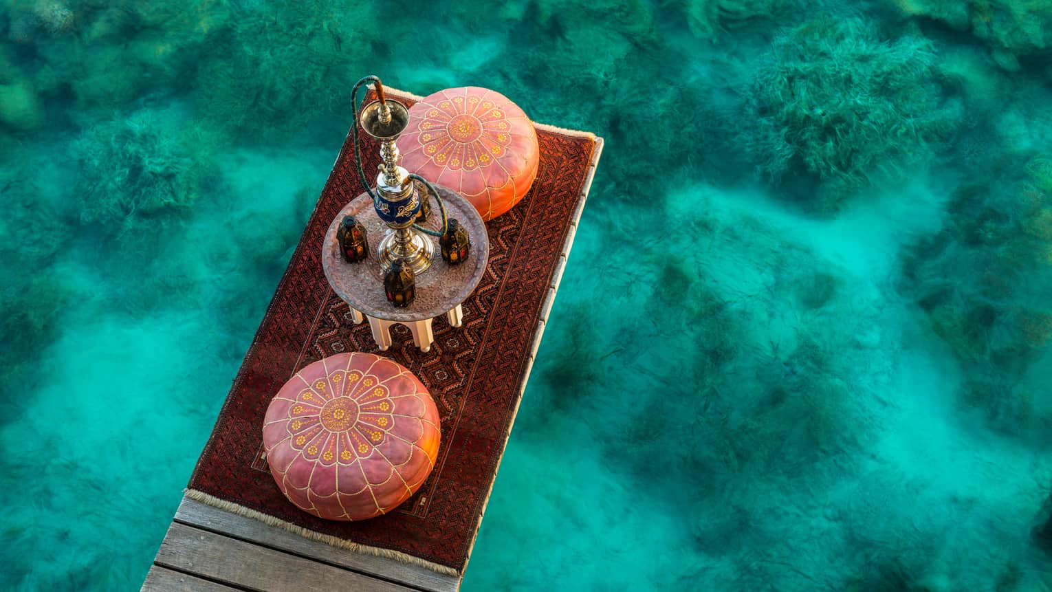 Aerial view of shisha pipe, two round cushions, red carpet on dock over turquoise lagoon