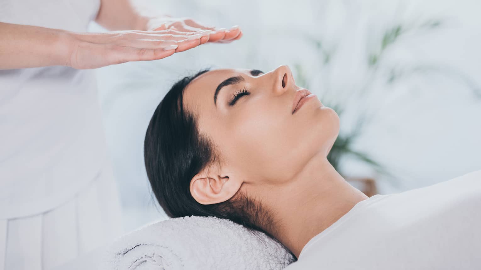 Woman with eyes closed lays on rolled up towel on spa table under therapists' hovering hands