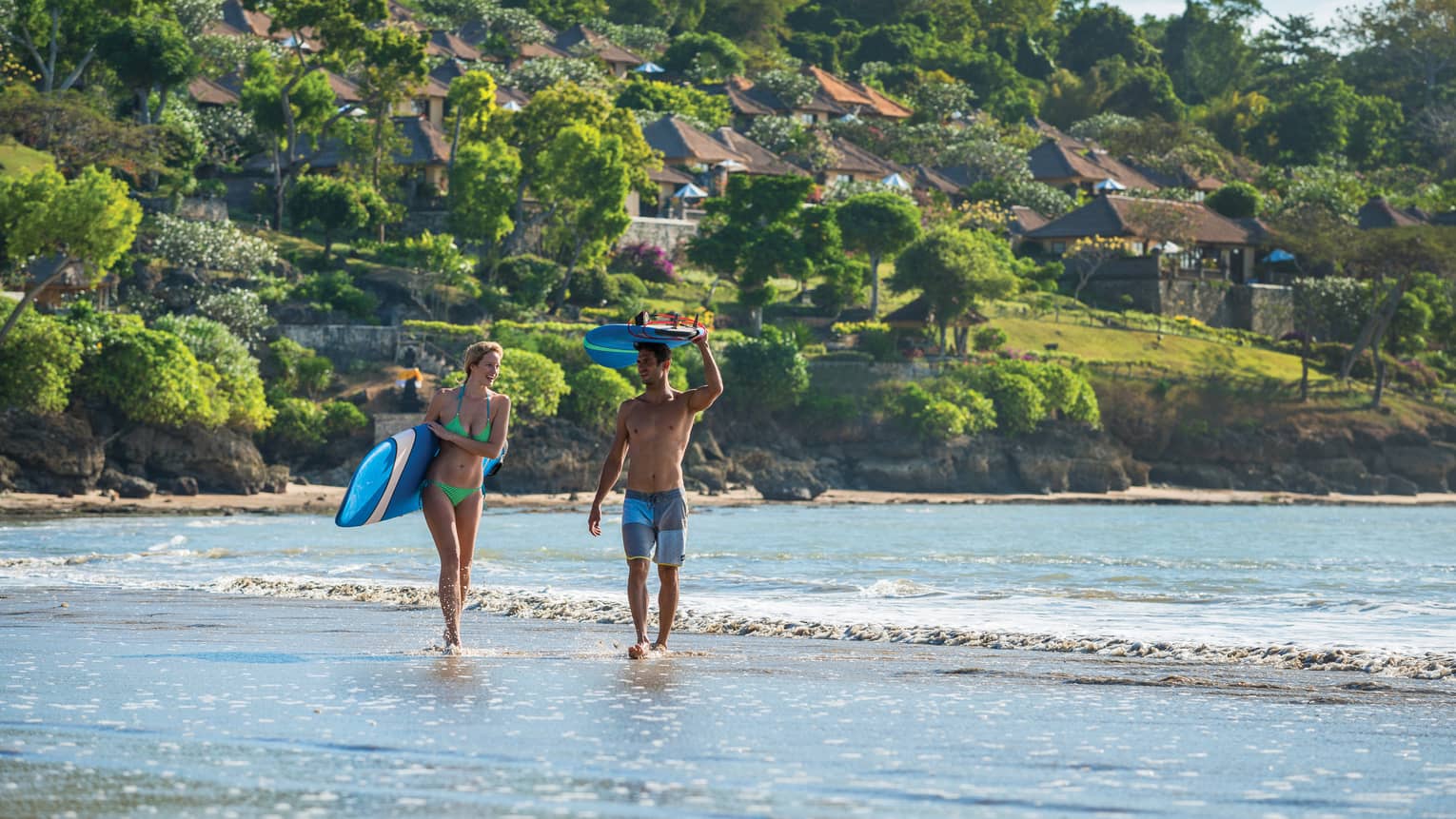 Man and woman in swimsuits walk on beach through surf, woman holds surfboard at side, man holds his above head