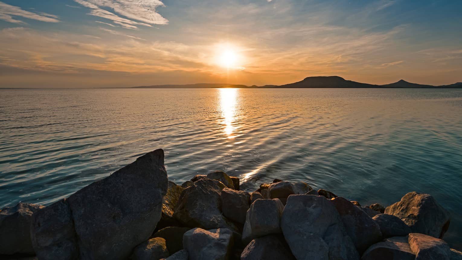 A jagged rocky shoreline looking out across the lake at hills being silhouetted by the setting sun.