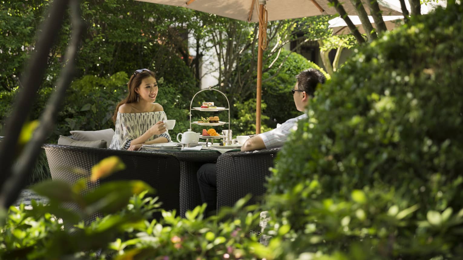 Woman and man drink tea near tray of sweets at table under shade of patio umbrella