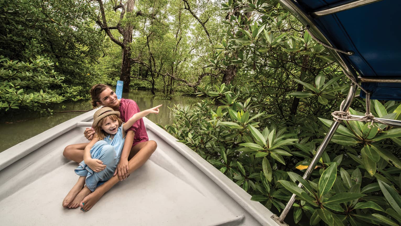A woman and child on the bow of a canopied riverboat amid lush, green mangrove trees; both gaze where the child is pointing.