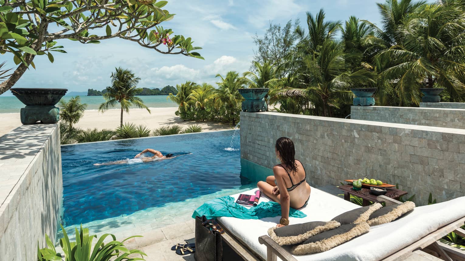 Back of woman in swimsuit on lounge chair, man swimming in pool below