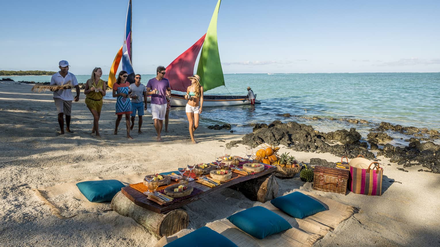Group of friends walk past colourful sailboats to beach lunch picnic