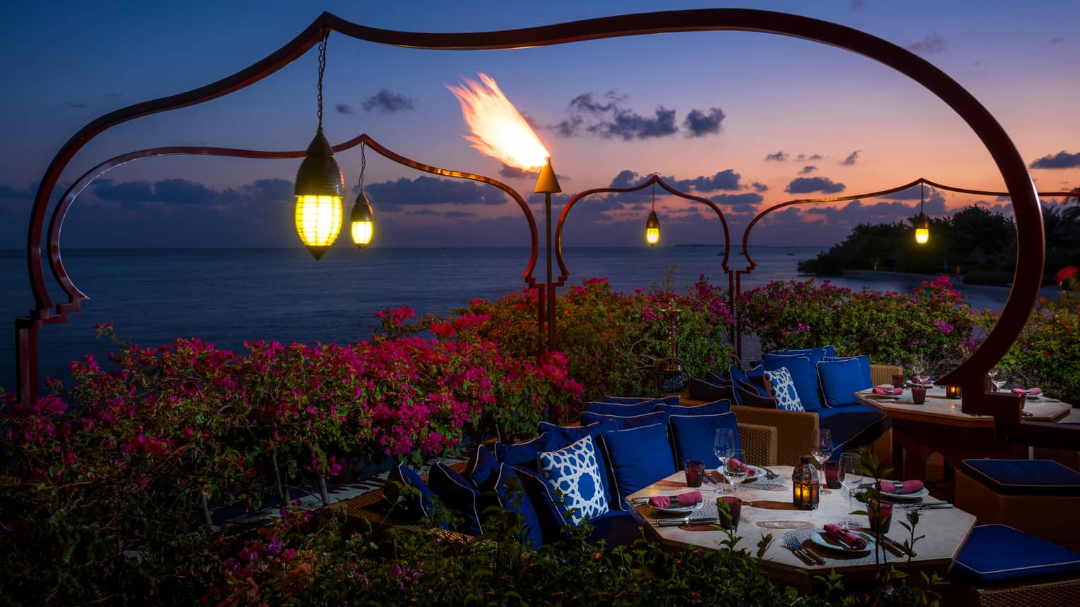 Lanterns hang over pink tropical gardens, booths with dining tables, plush blue pillows at night
