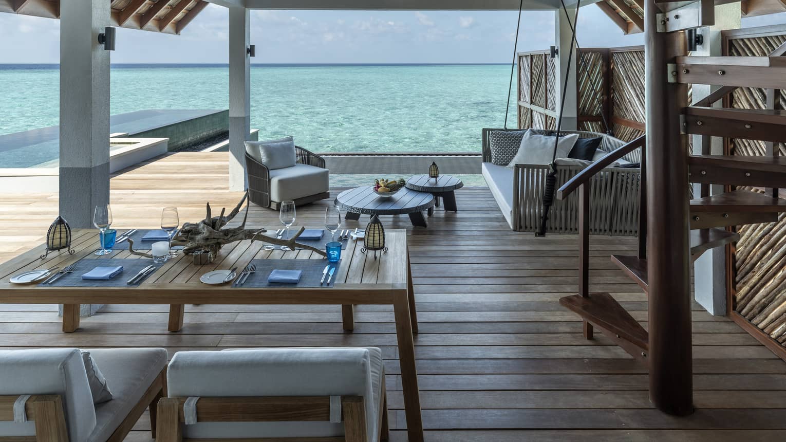 Outdoor terrace of an over-water villa, featuring wooden floors, table and chairs, lounge seating, staircase