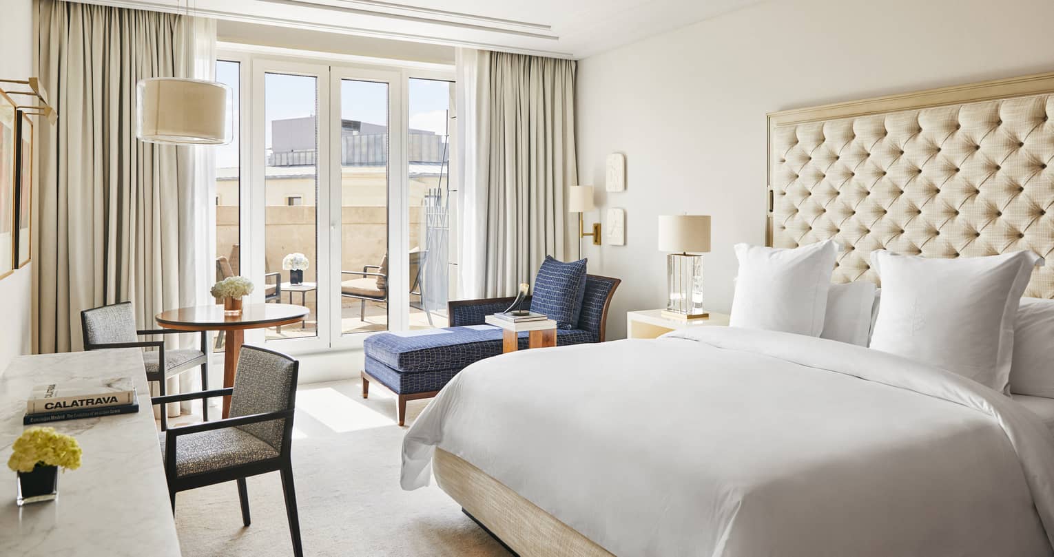 Luxury hotel bedroom with a king bed, tufted headboard, blue chaise lounge, round table and desk with chairs and French doors leading to a terrace