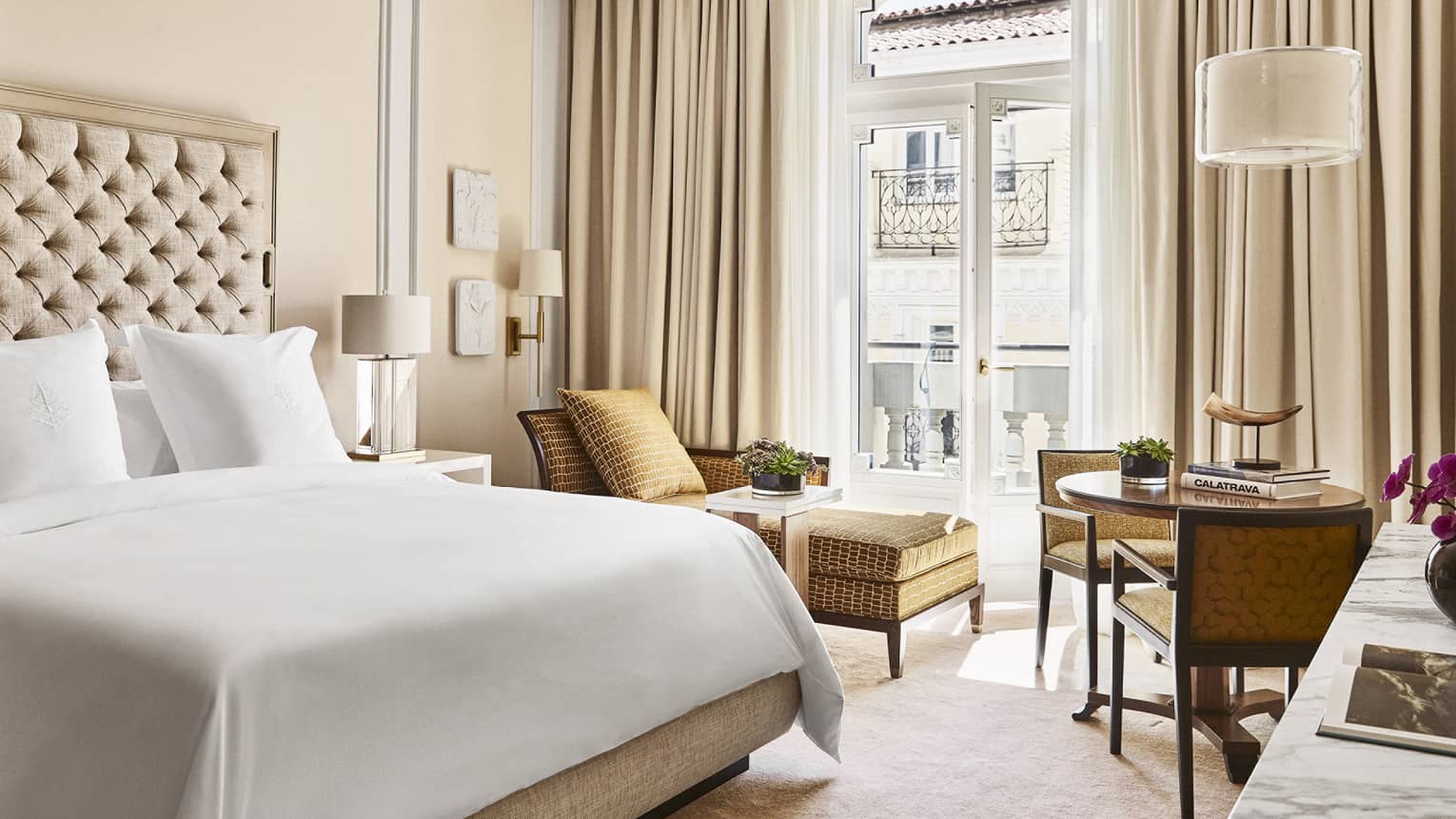 Luxury hotel bedroom with king bed, tufted headboard, table with books and horn sculpture, tan chairs and chaise lounge and doors opening to a Juliet balcony