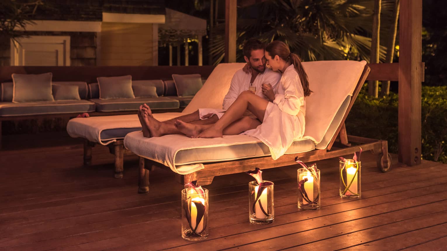 Man and woman in bathrobes relax on lounge chairs, glowing candles on deck at night