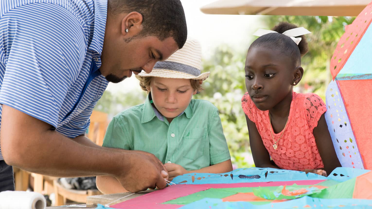 Man ties string onto colourful paper kite at table as two children watch