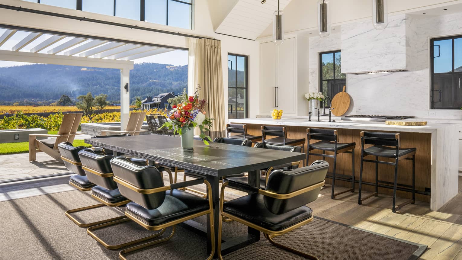 Kitchen and dining area with dark wood table, black leather chairs, marble island and open sliding doors leading to patio with vineyard and mountain views