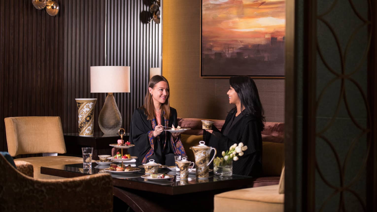 Two women in thin black robes sit on sofa, enjoy high tea at table