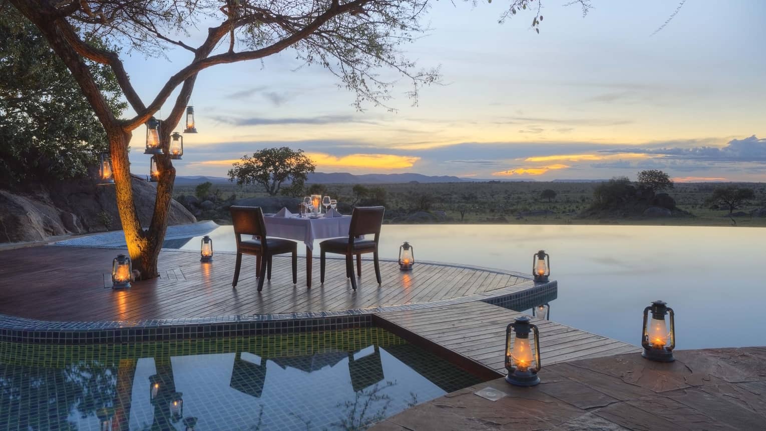 Private dining table on platform lined with lanterns over infinity swimming pool at desk