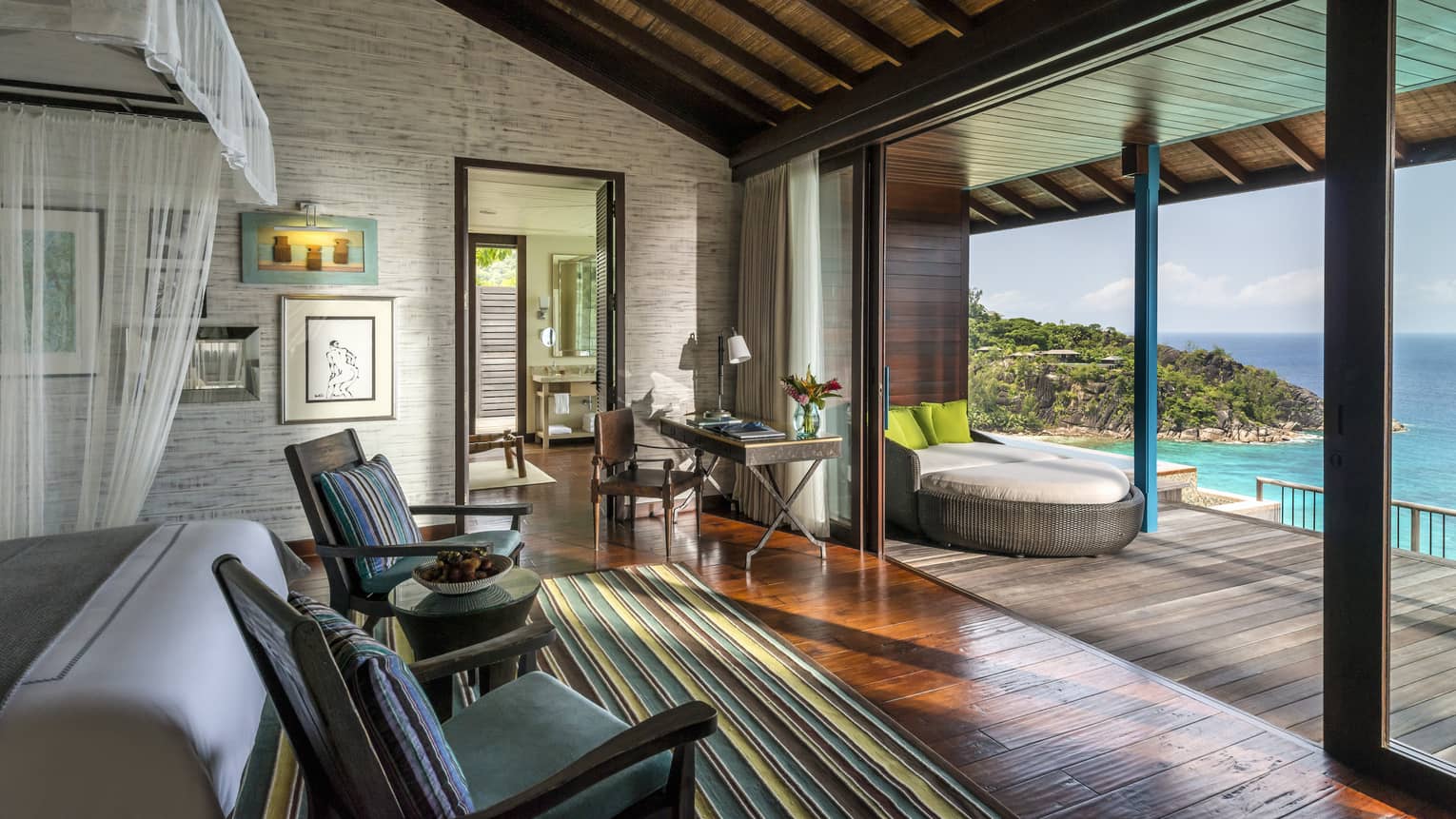 Villa with bed, two chairs, opening up to patio with lounge chair and ocean view 