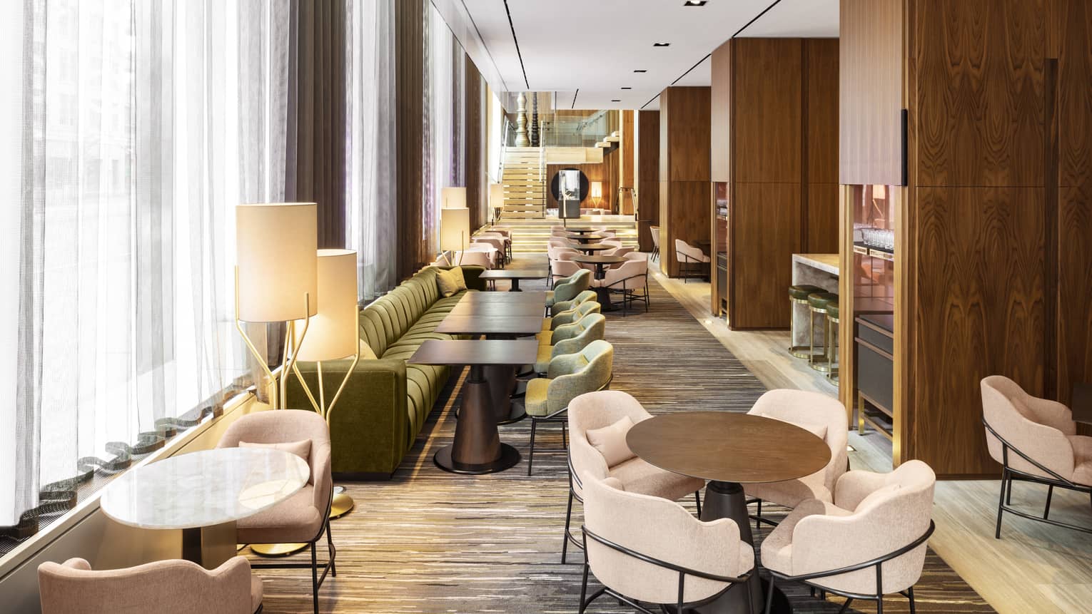 A long room lined with upholstered sofas and chairs, wood tables and windows 
