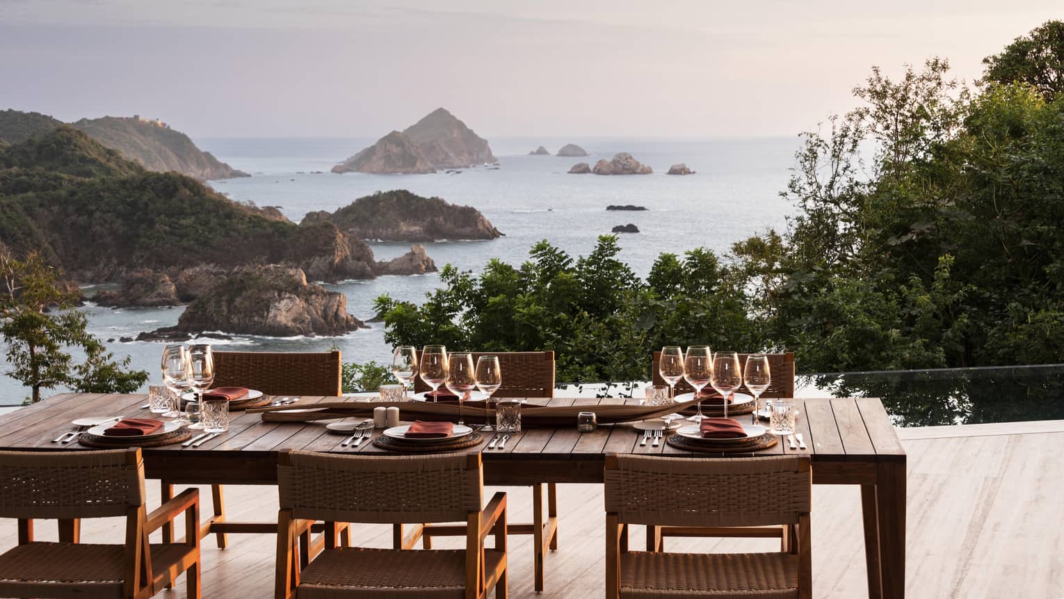 A beautifully set outdoor dining table with glassware and plates, overlooking a breathtaking ocean view with rocky islands and lush greenery at sunset