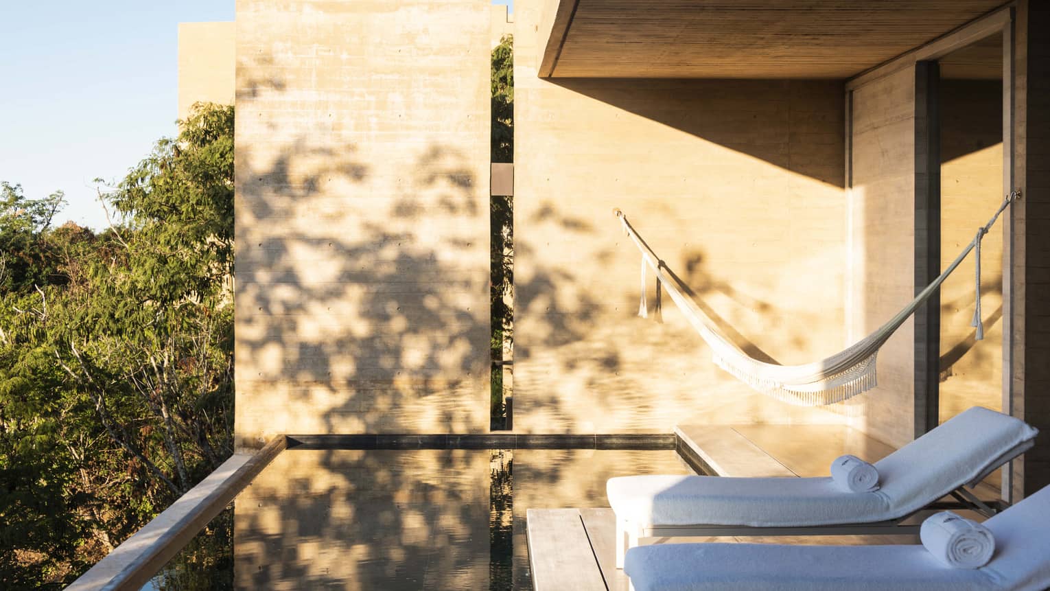 Sunlit outdoor terrace with lounge chairs and a hammock, overlooking a small infinity pool. The space is surrounded by trees, with natural wood walls casting soft shadows.