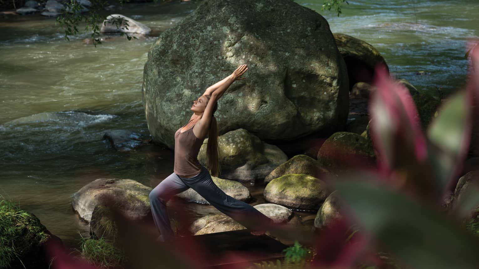 A guest practices yoga on the banks of a forest river; sunlight dapples the flowing water and the large, mossy boulders.