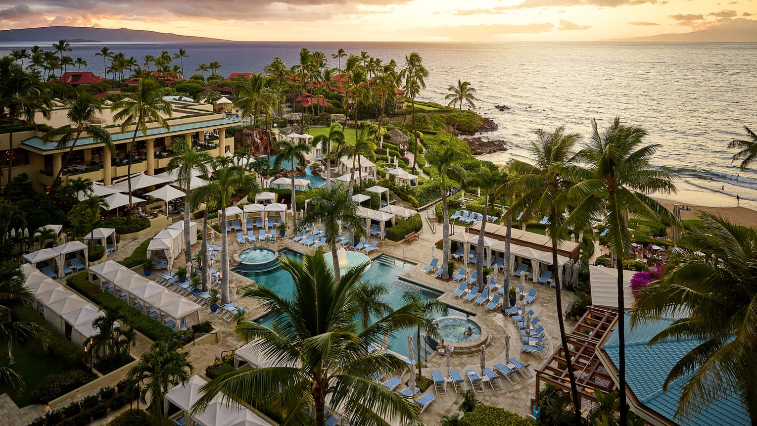 Tropical luxury resort pool lined with blue lounge chairs and palm trees, set next to ocean at sunset