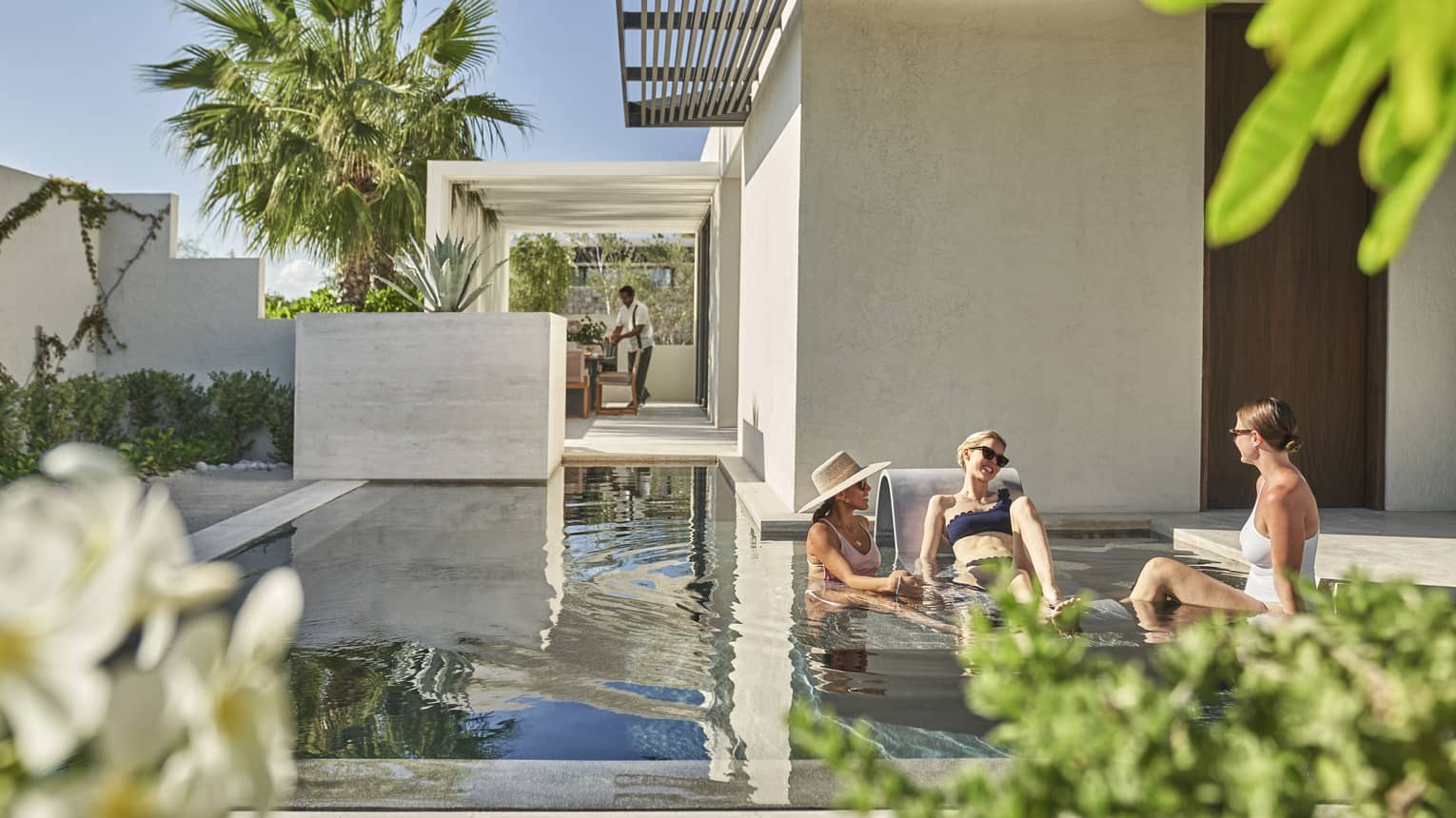Three people sitting on a shallow pool outside, palm trees and blue skies can be seen behind them.