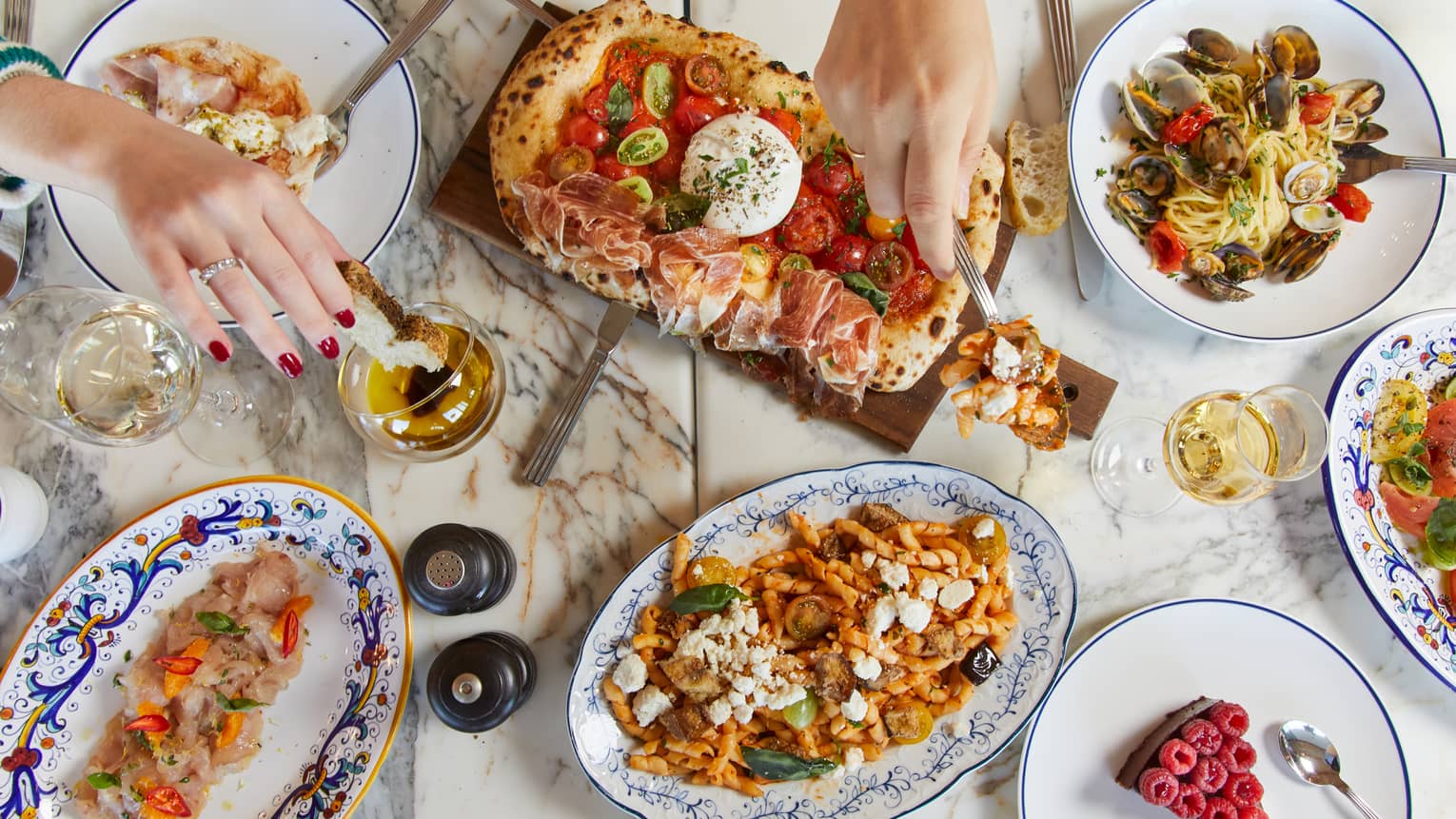 A marble table features white wine and an array of colourful Mediterranean delicacies, hands reaching in to sample the food.