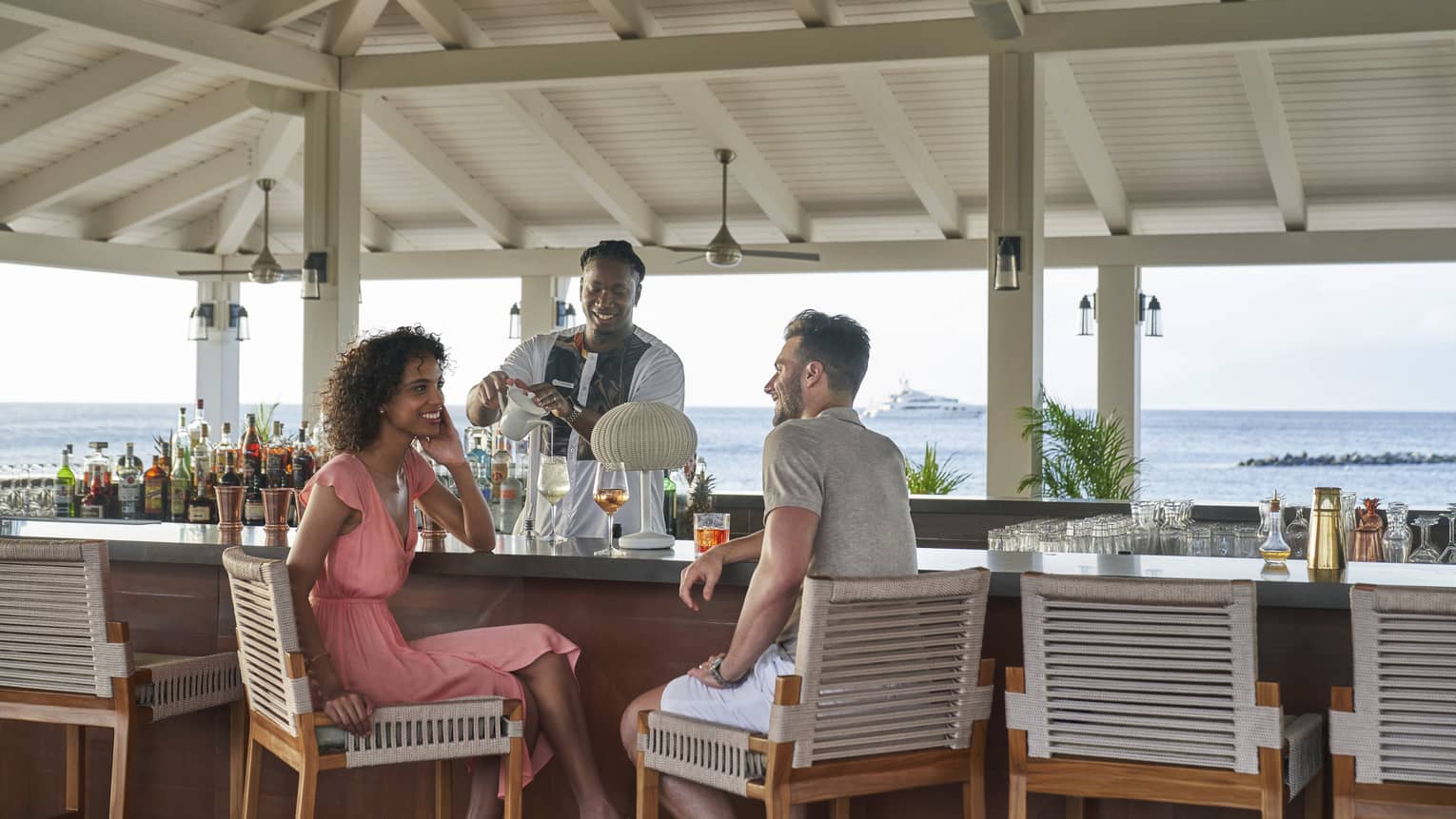 A man and woman talking at a bar outside while the bartender makes a drink.