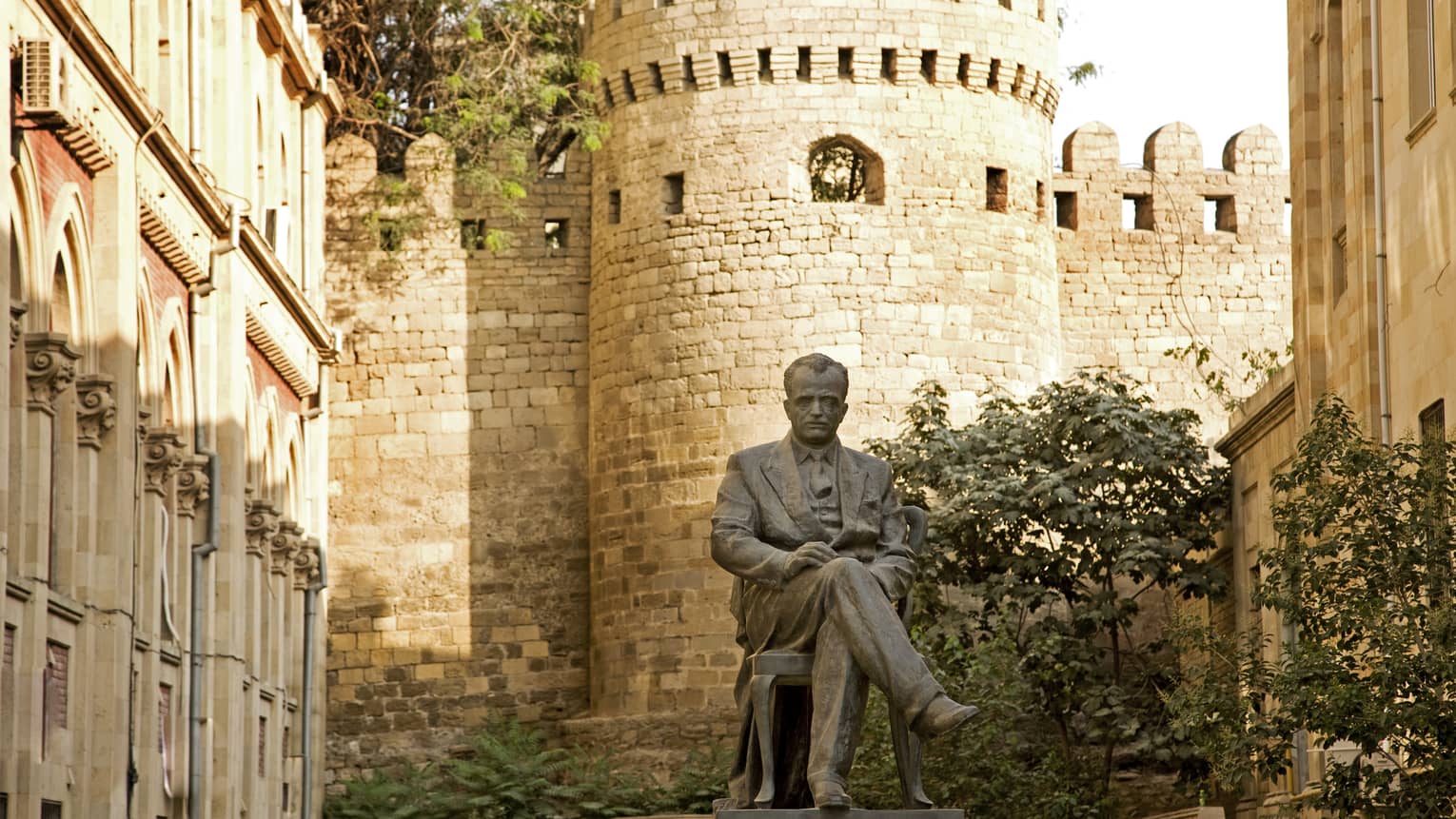 Surrounded by stone fortress, the statute of a man seated on a pedestal in full suit and tie, legs crossed, hands on his lap.