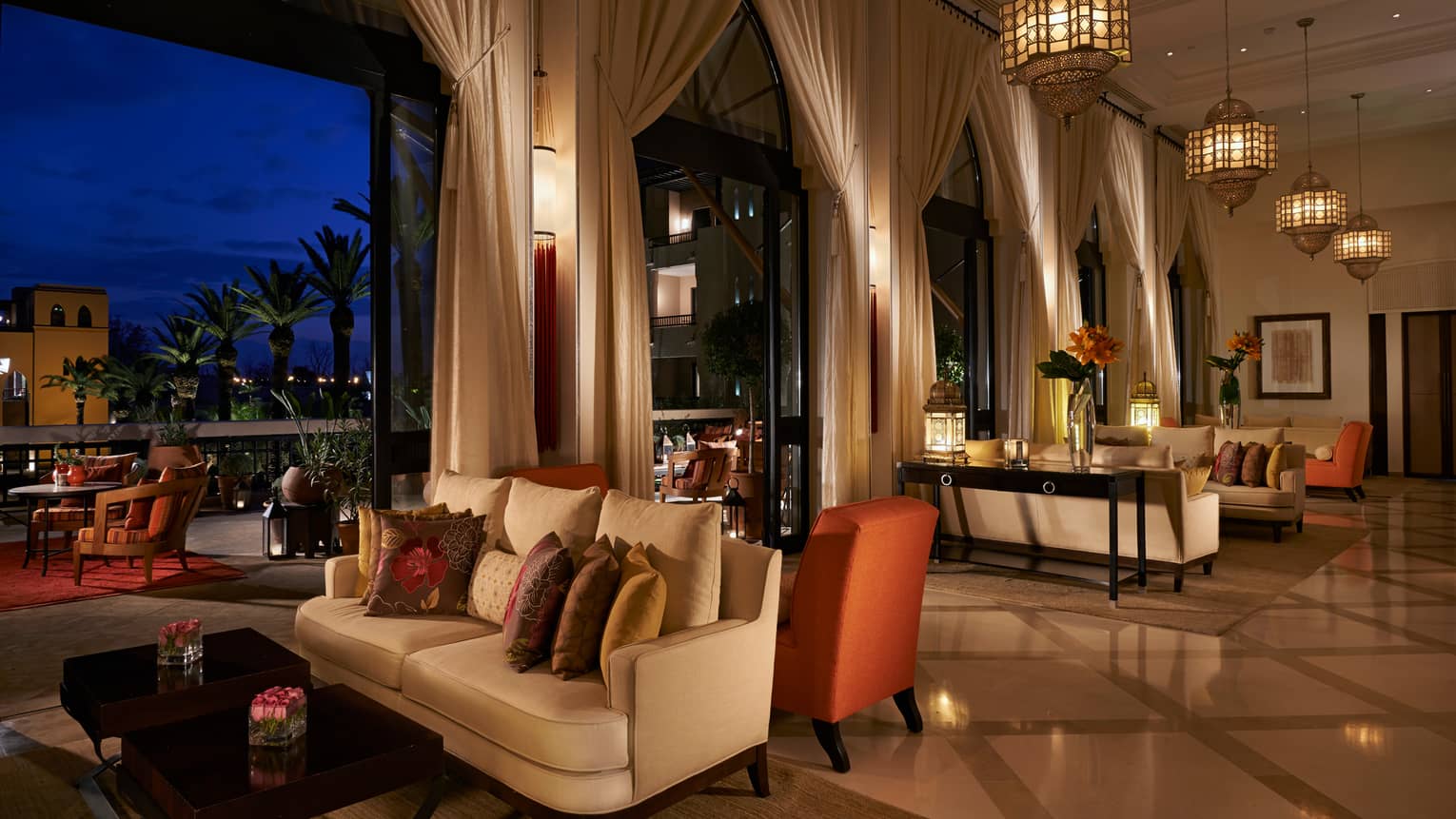 Inara Lounge indoor seating area at night with small white sofas, orange chairs, tall open-air windows with white curtains